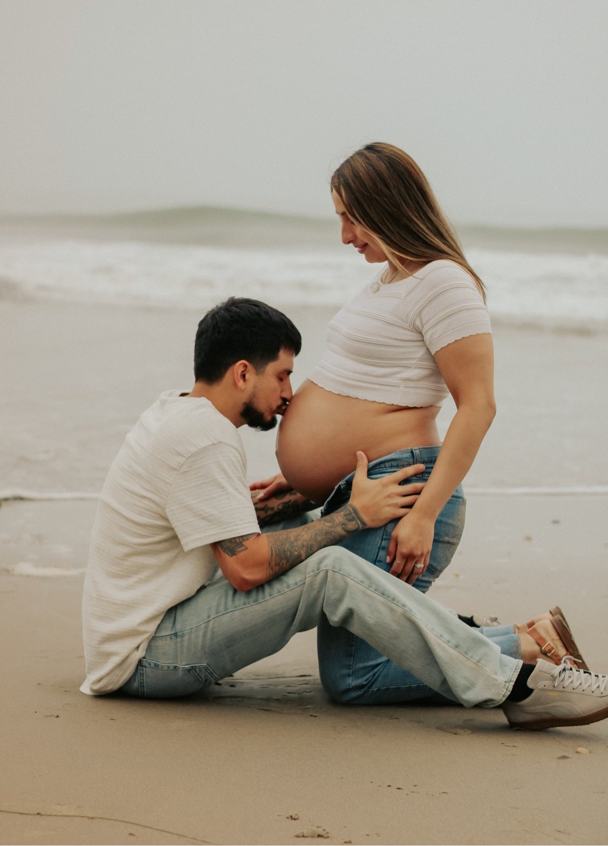 Partner kneeling on the beach kissing a pregnant belly as waves roll in