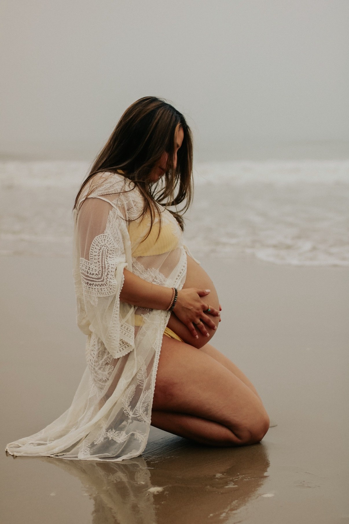 Expecting mother kneeling in the surf in a white lace cover-up, hands on her belly