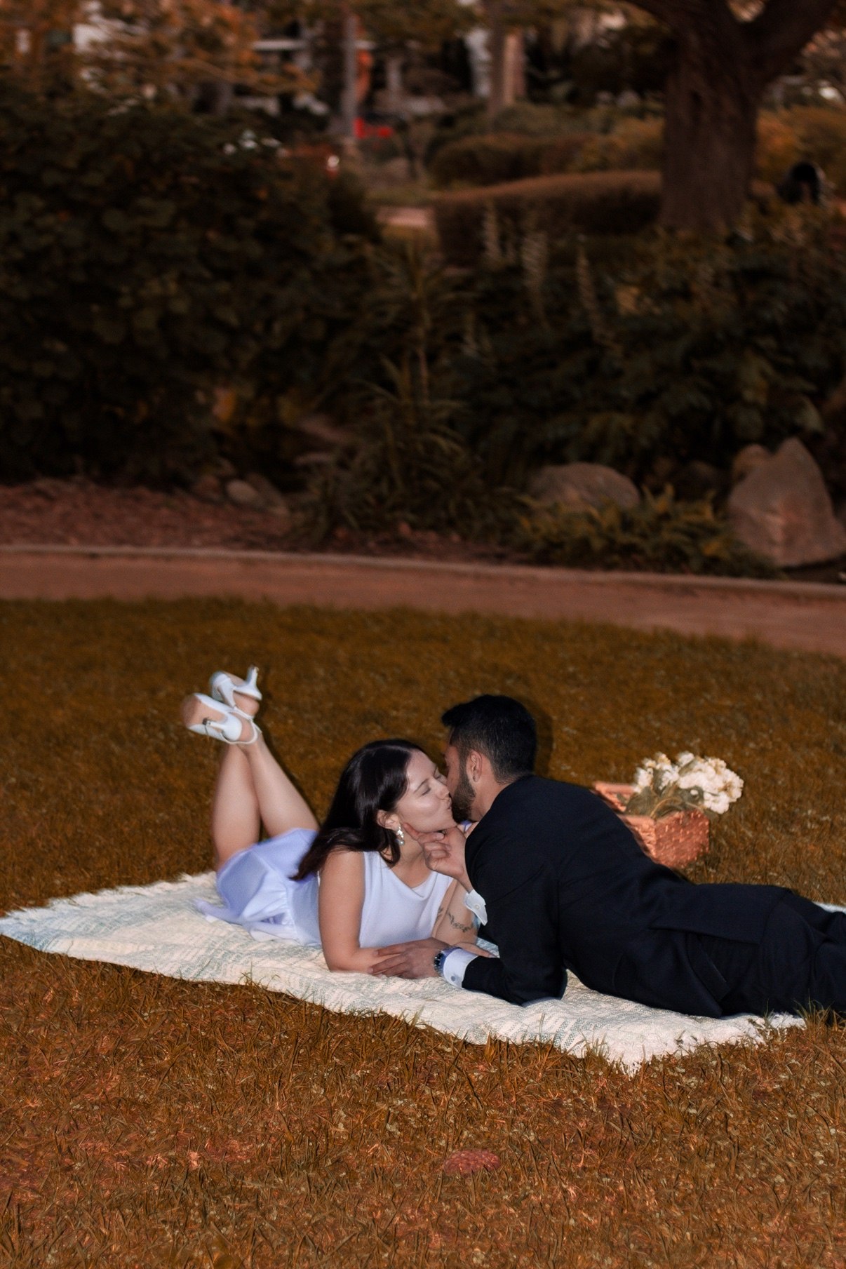 Couple lying on a picnic blanket on the grass, kissing, with a basket of white flowers beside them