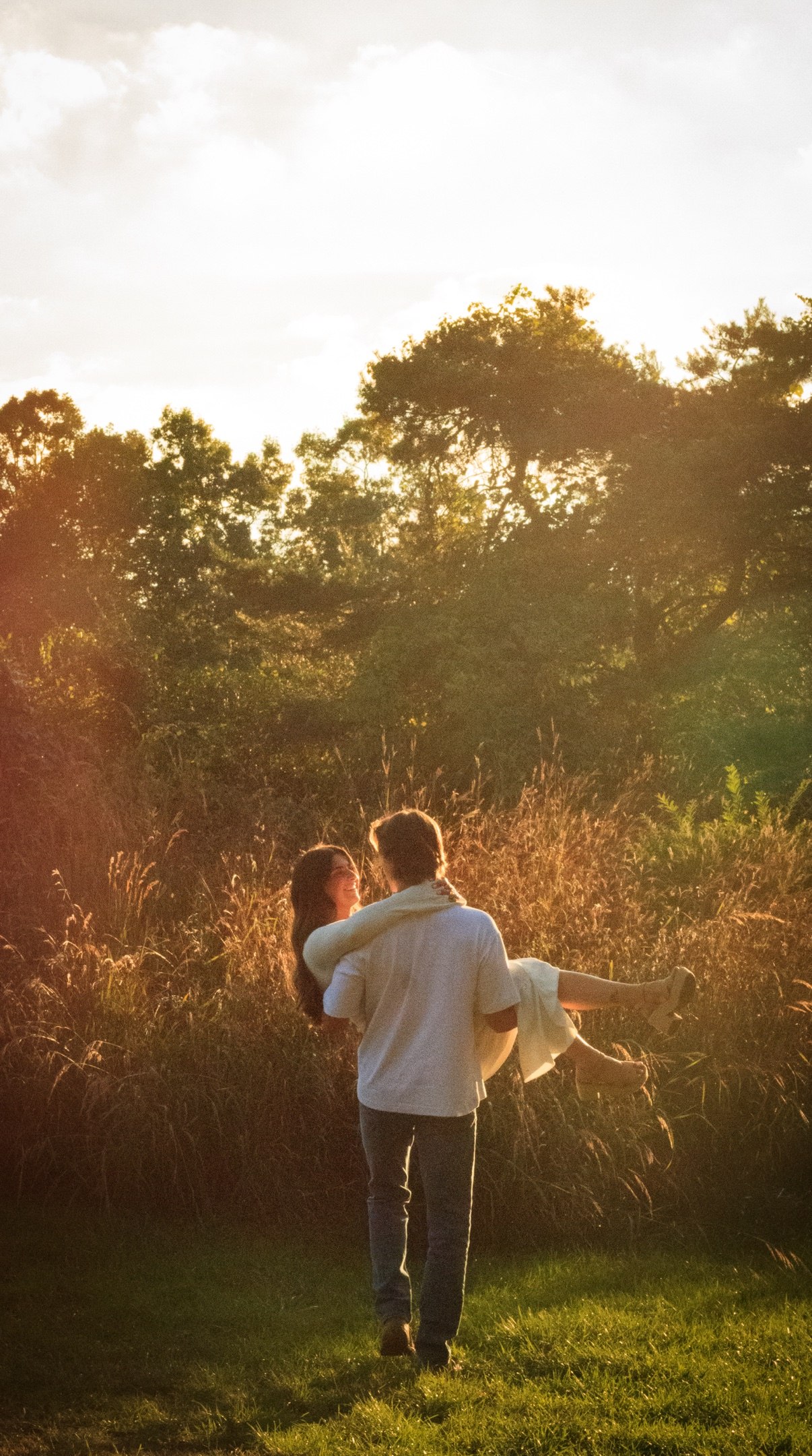 Man carrying his partner in his arms through tall grass at golden hour, both laughing