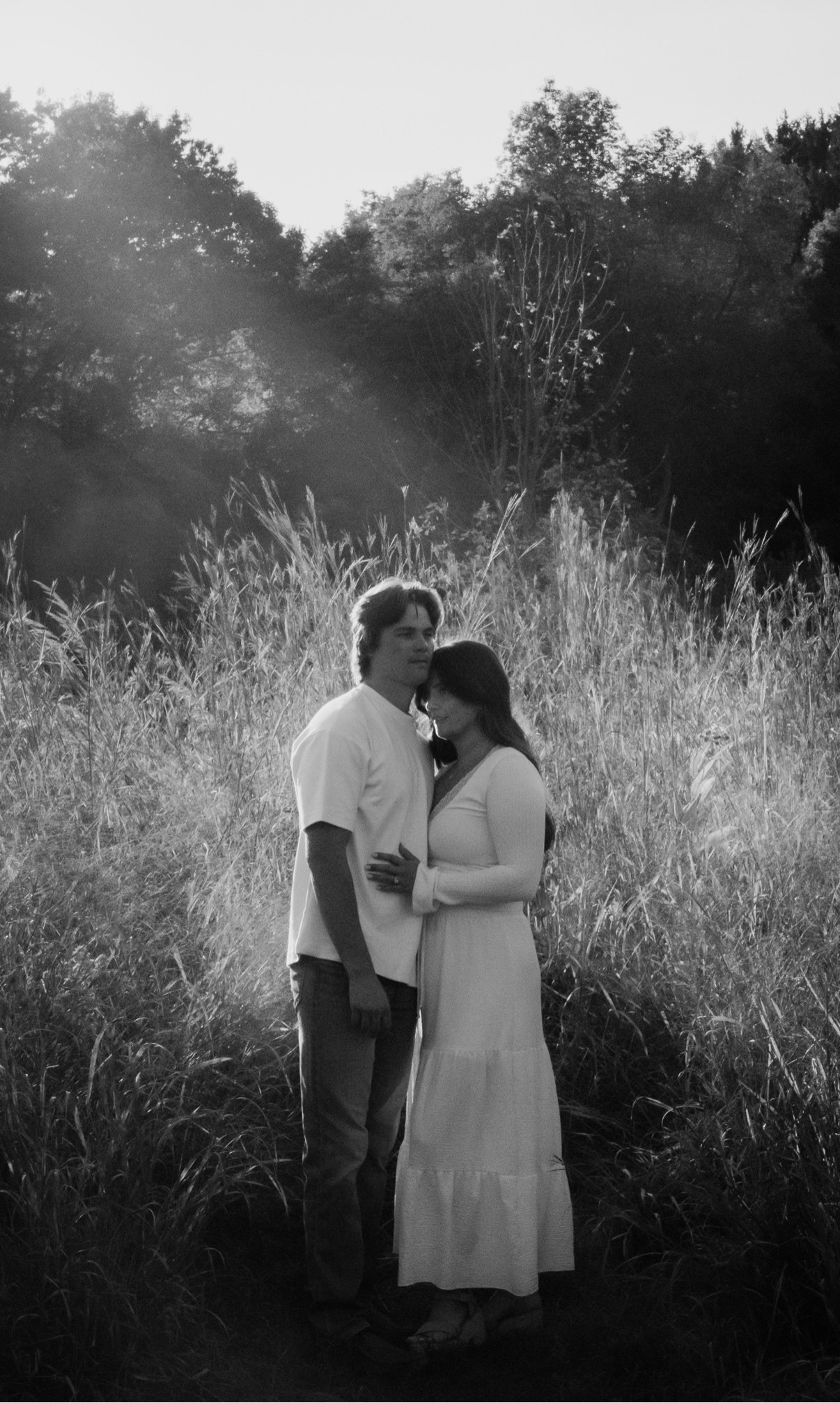Black-and-white photo of a couple embracing in tall grass, her head resting against his chest