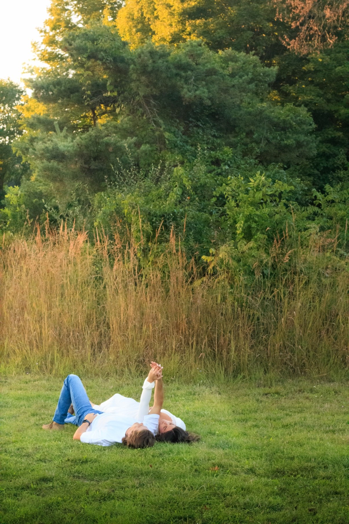 Couple lying side by side on the grass holding hands up toward the sky, trees and tall grass behind them