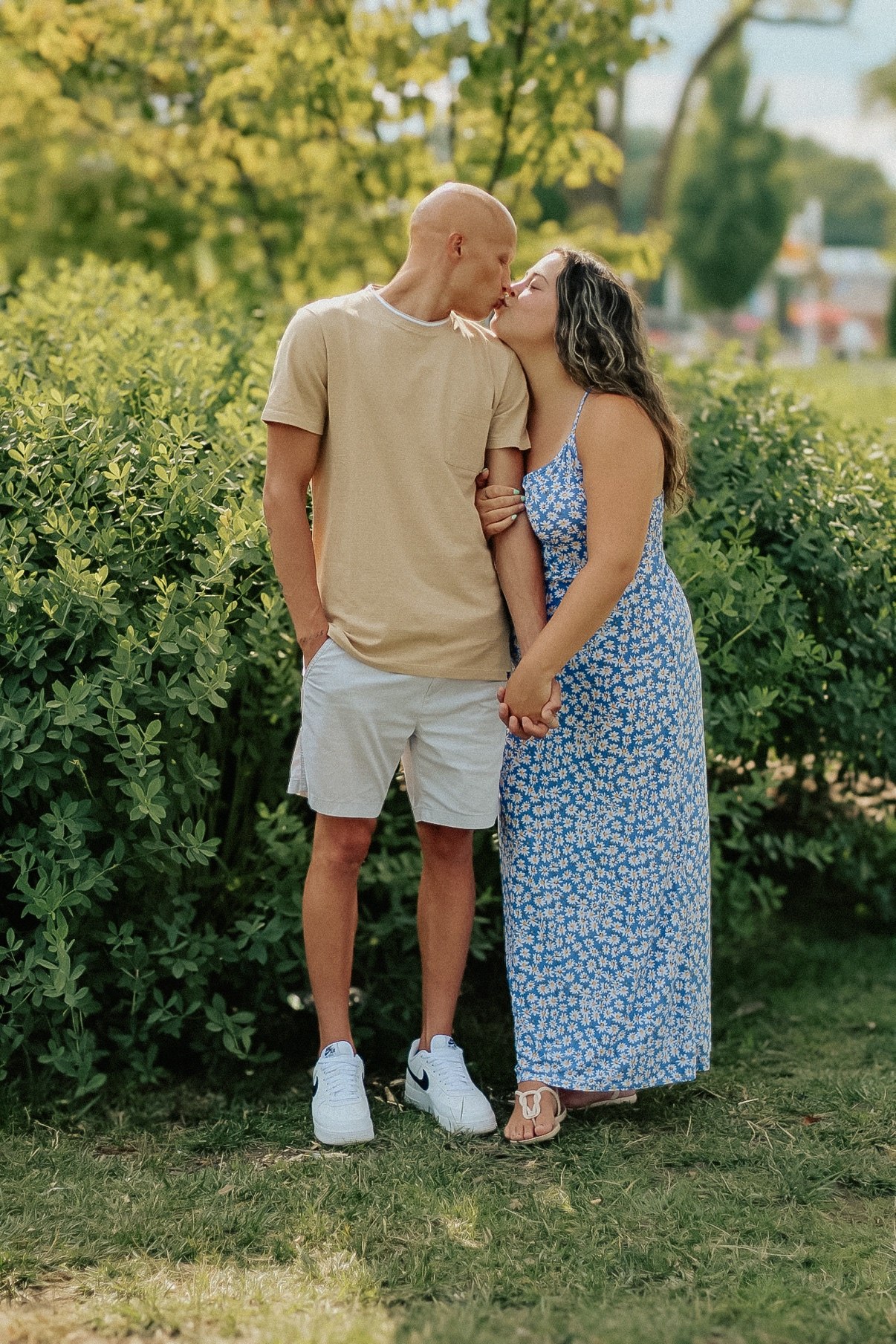 Couple kissing and holding hands in front of green hedges, the woman in a blue floral maxi dress