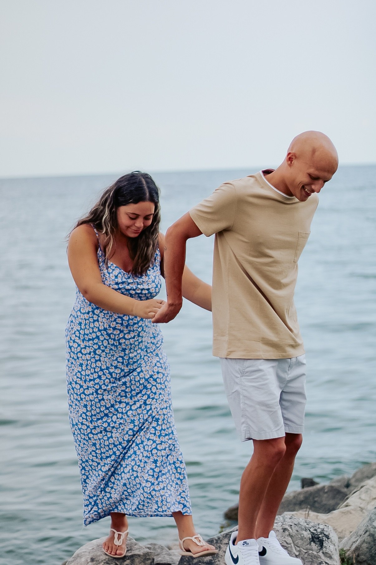 Couple laughing and holding hands while stepping across rocks at the edge of the water